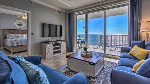 A spacious living room with a large sofa, a coffee table, and a television. There is a balcony with a view of the city.