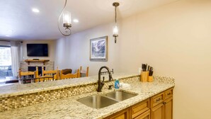 Una cocina con encimeras de granito, armarios de madera, un lavamanos doble y un comedor con sillas y una mesa.
