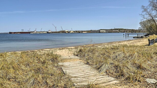 Una playa arenosa con un paseo marítimo de madera que conduce al agua, un área industrial distante con grúas, y un cielo azul despejado.