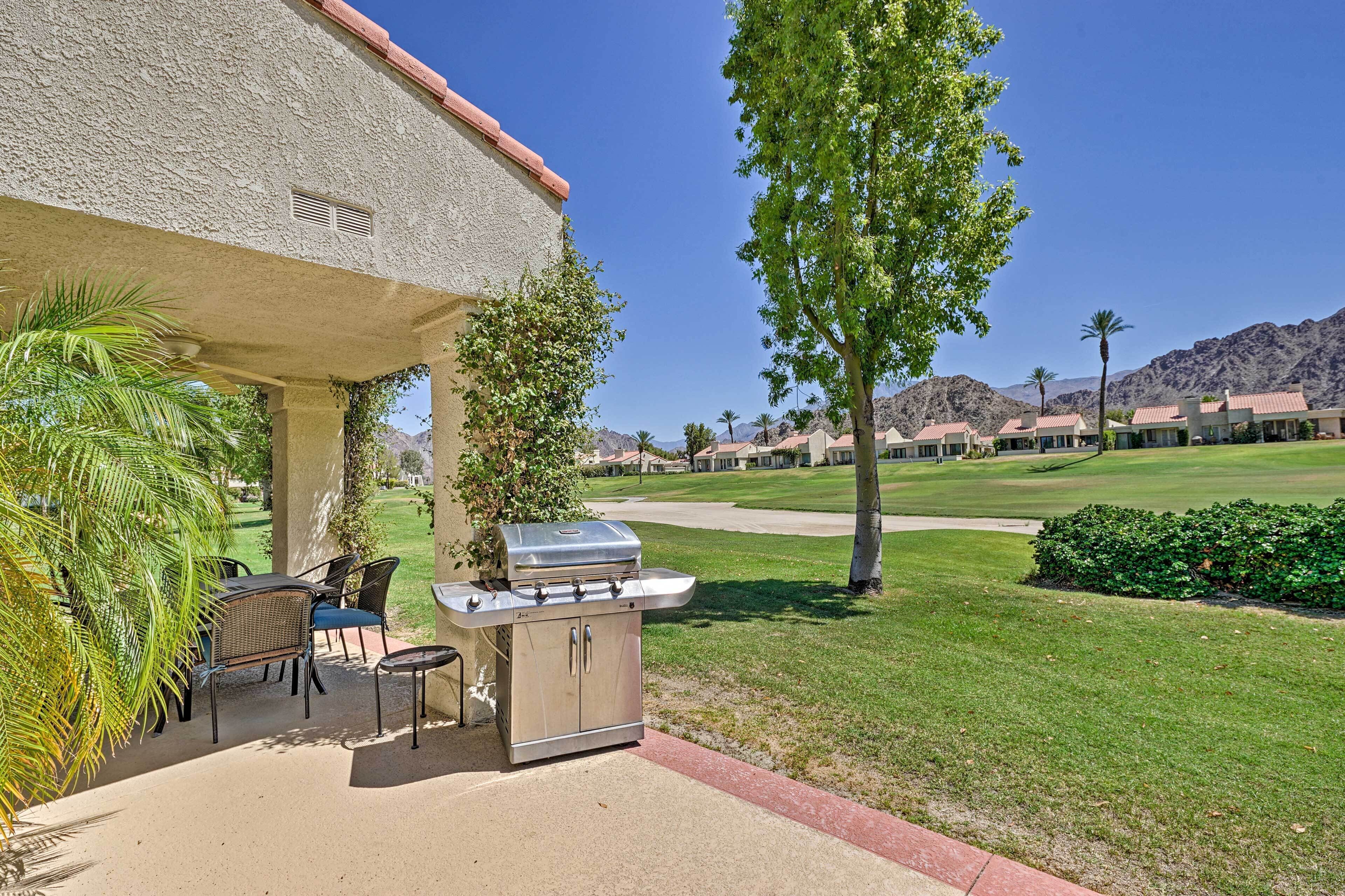 An outdoor patio with a grill, chairs, and a view of a golf course and mountains.