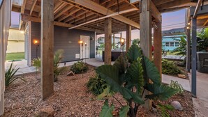 A covered outdoor patio with wooden beams, a gray building in the background, and a garden area with large plants and mulch. - Walk to Sea Isle Beach: Updated Group Getaway! (Galveston)