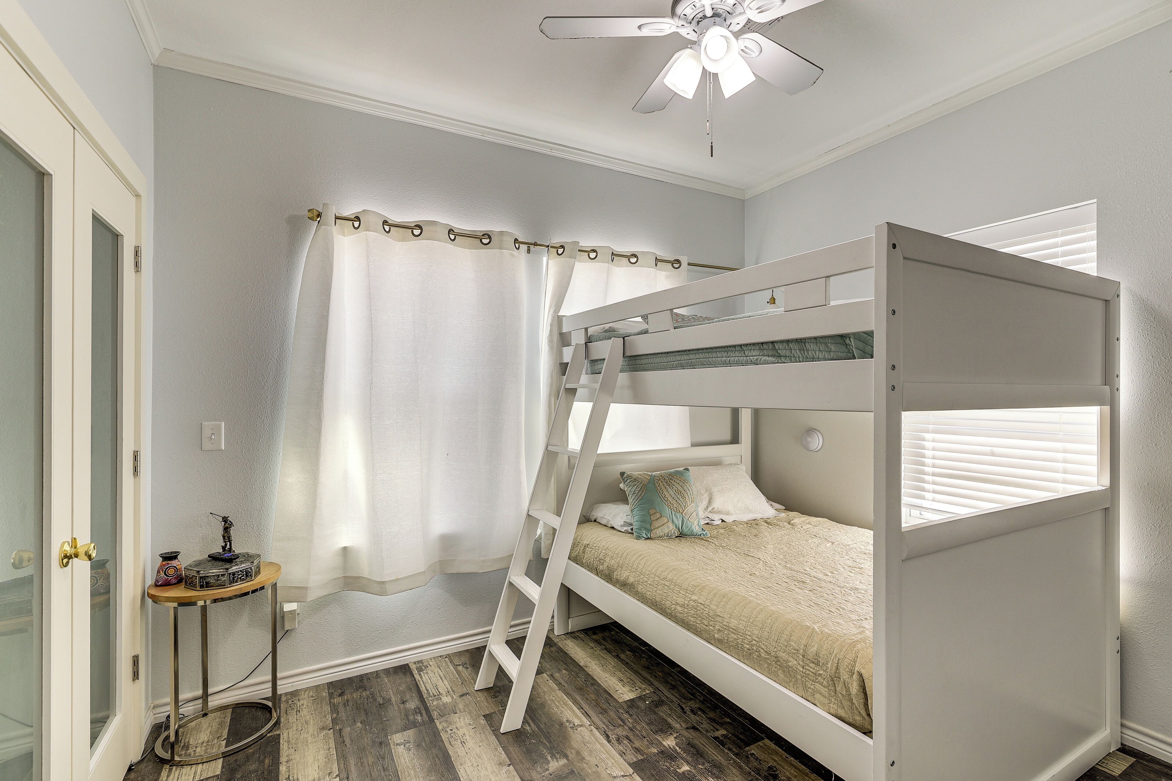 Une chambre avec un lit superposé, une échelle, une petite table agrémentée d’un objet décoratif et une fenêtre avec des stores.