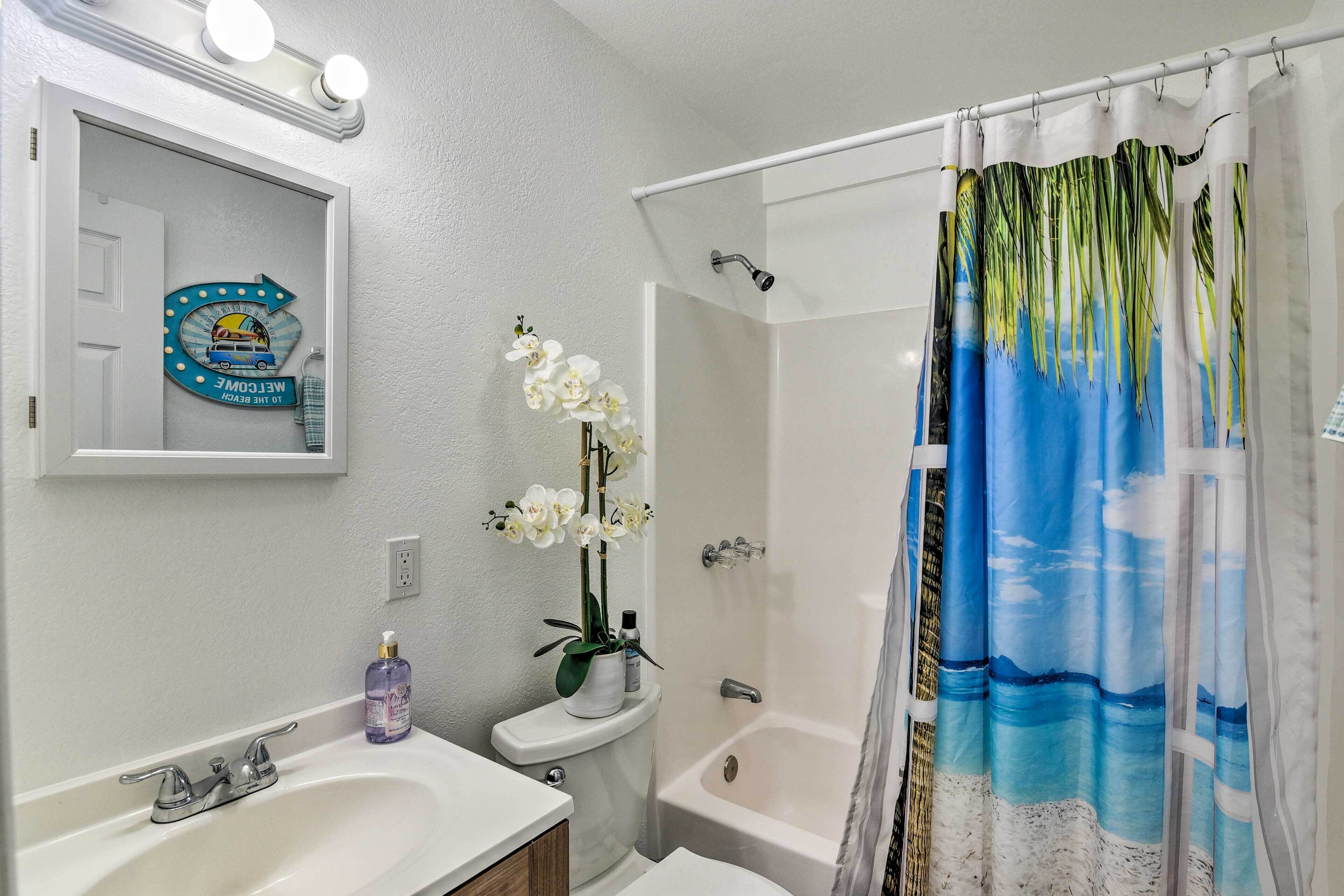 A bathroom with a white sink, a toilet, a shower, and a bathtub. There is a shower curtain with a tropical design, and a small potted orchid on the toilet.