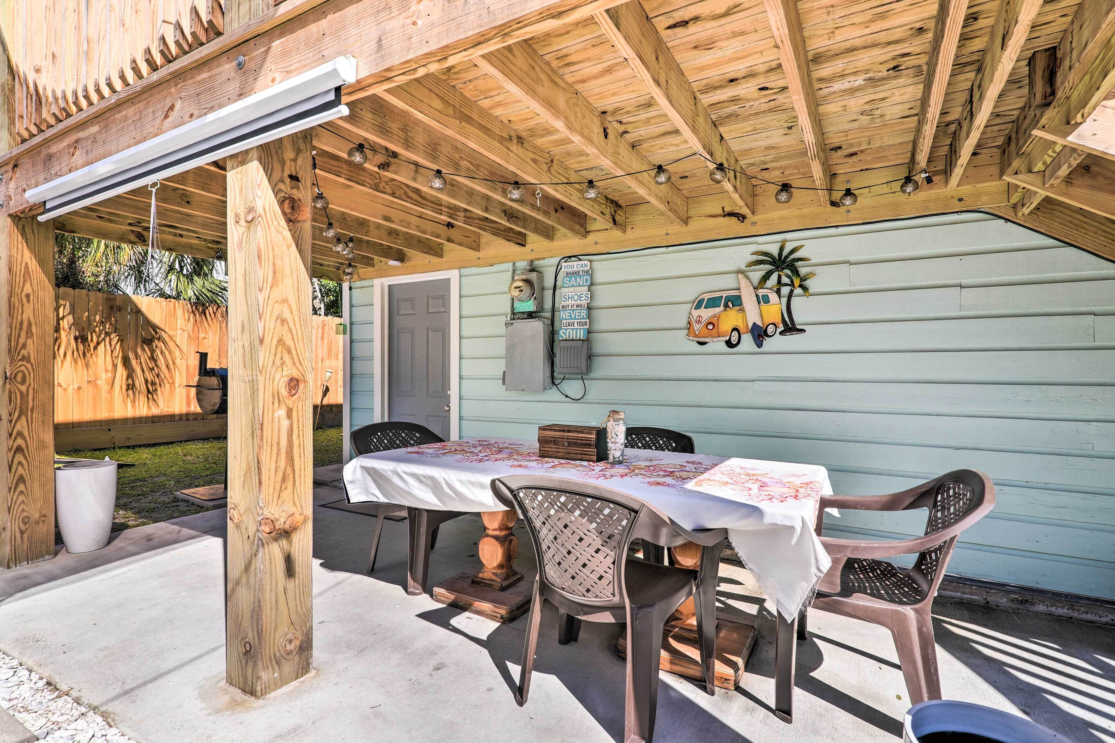 A covered patio with a table and chairs, wooden beams, a string of lights, and a wall with a van and palm tree decoration.