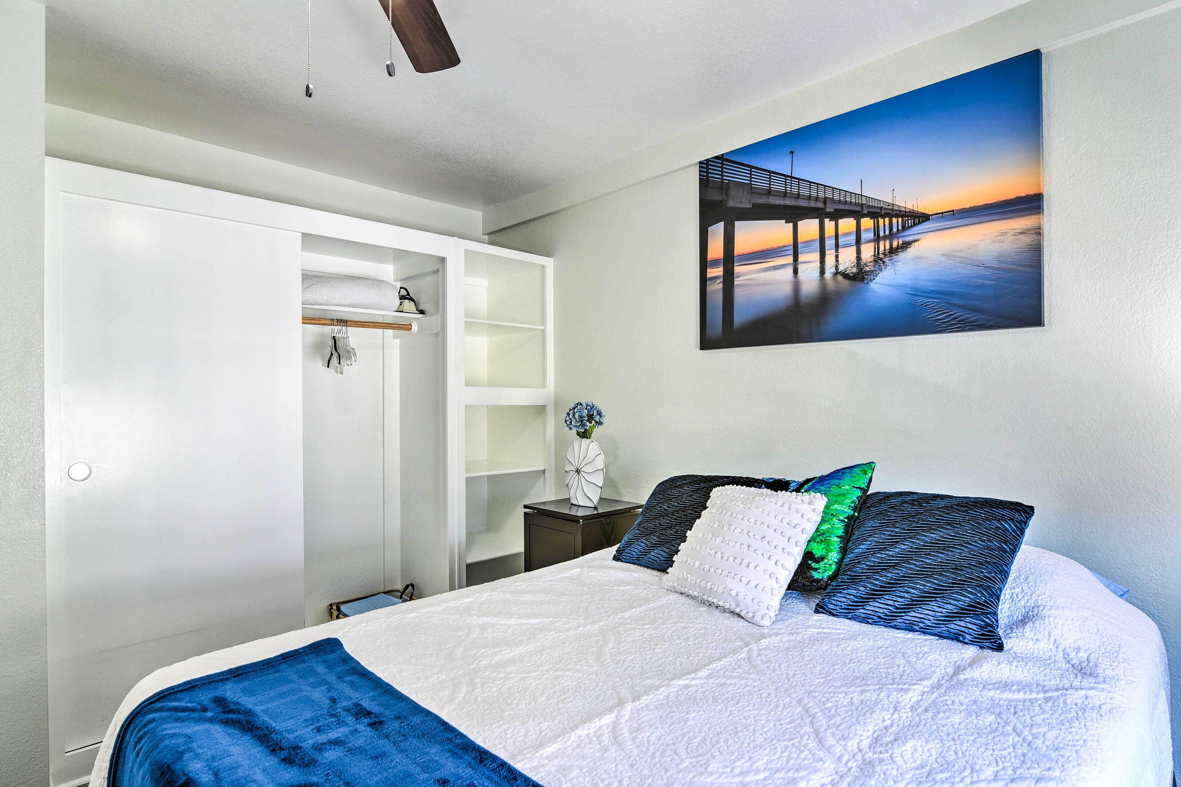 Une chambre à coucher comprenant un lit, une table de chevet, une commode, un miroir et un tableau représentant une jetée au coucher du soleil.