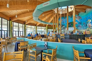 A restaurant with wooden ceiling beams, large windows, and a mural of marine life. Tables and chairs are arranged for dining.