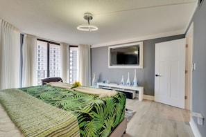 A modern hotel room with a large bed, a flat-screen TV mounted above a white console, and a large window with curtains.