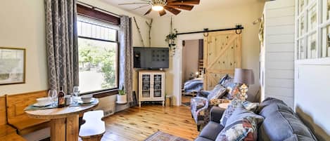 Un salón acogedor con piso de madera, un sofá con cojines estampados, una mesa de madera con un banco, un ventilador de techo y un televisor sobre un mueble.