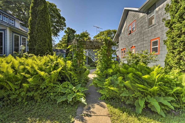 A pathway flanked by lush greenery and a wooden pergola, leading to a house with a grey facade and red-framed windows.