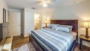 Un dormitorio con cama, cabecero de madera, dos mesitas de noche con lámparas, un ventilador de techo y una puerta que da a otra habitación.