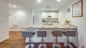 Una cocina moderna con armarios blancos, encimera de granito y microondas integrado. Hay tres taburetes de bar y un reloj en la pared.