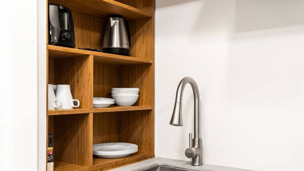 Une cuisine avec un évier en acier inoxydable, des étagères en bois où sont posées des tasses, des assiettes blanches et une machine à café.