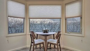 A corner room with a round table and two chairs, large windows with blinds, and a view of bare trees. - Cozy Anchorage Retreat - Near Hiking & Dtwn! (Anchorage)