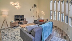 A spacious living room with a large flat-screen TV, a sofa, and a coffee table. There are multiple windows providing natural light and a view of the outside. - Cozy Anchorage Retreat - Near Hiking & Dtwn! (Anchorage)