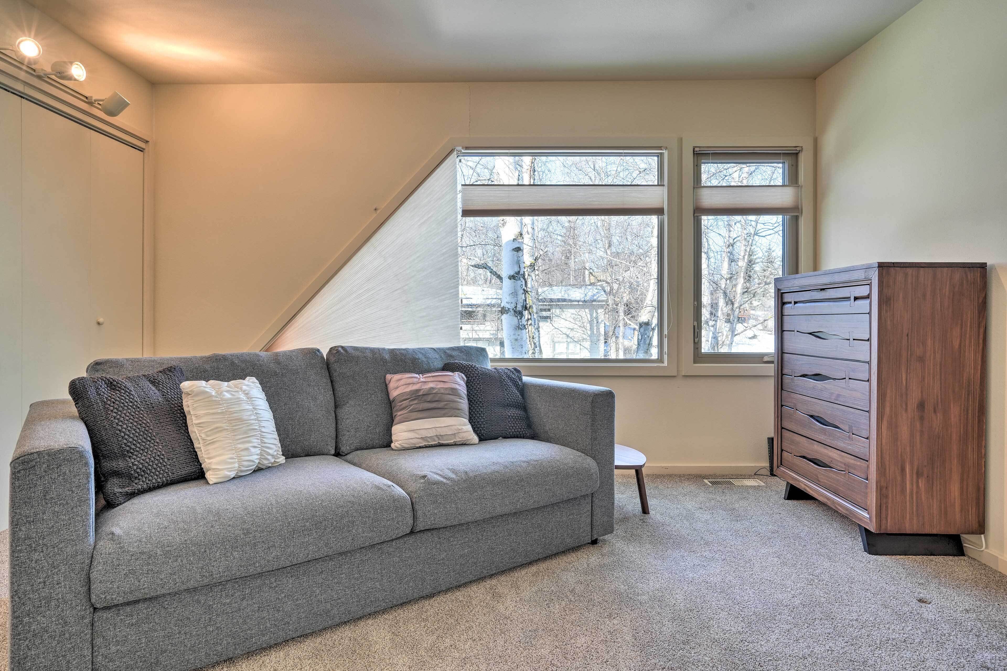 A room with a grey sofa, a wooden cabinet, and a window with a view of trees.
