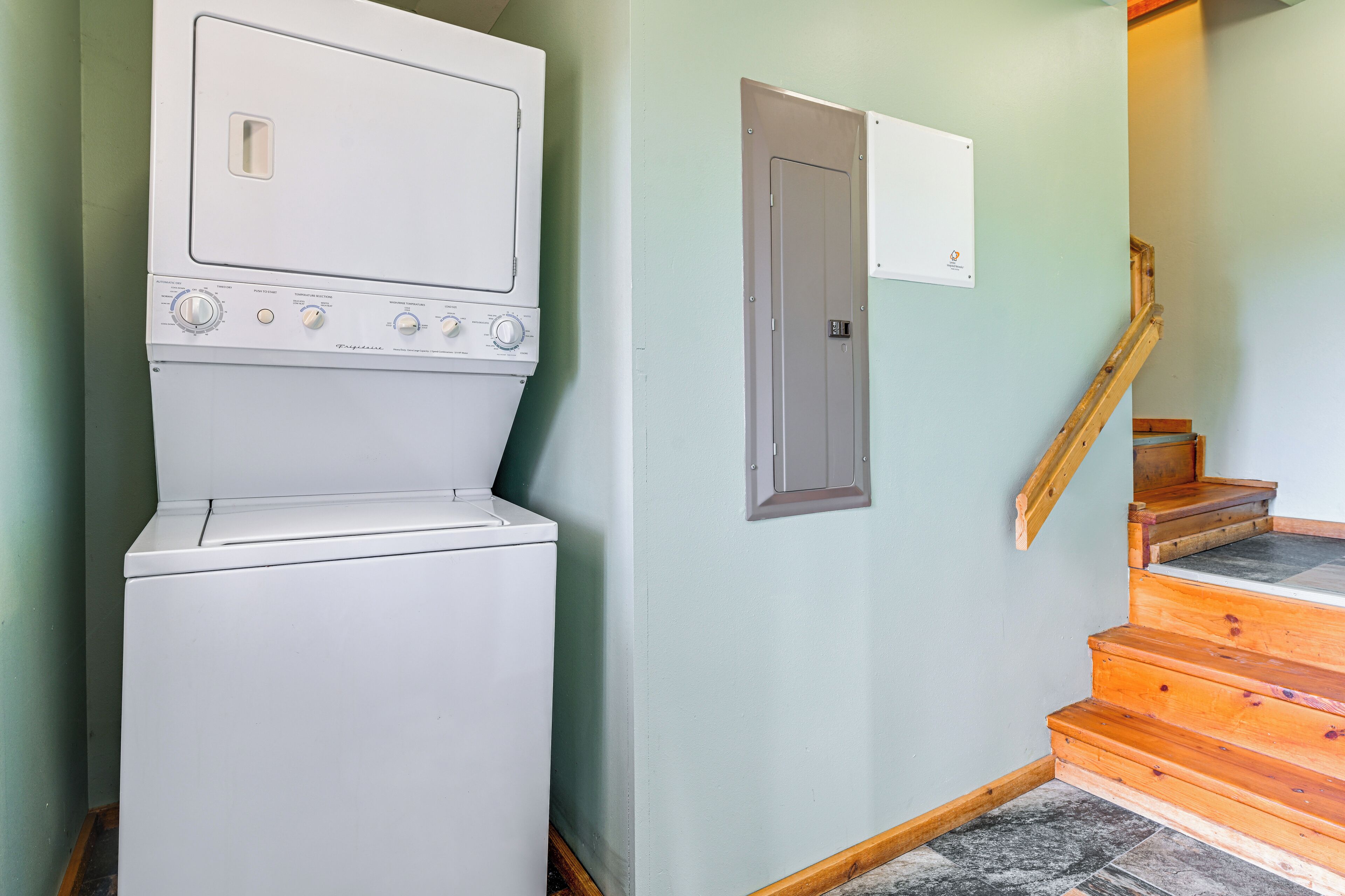 A laundry room with a stacked washer and dryer, a door with a label, and a wooden staircase.