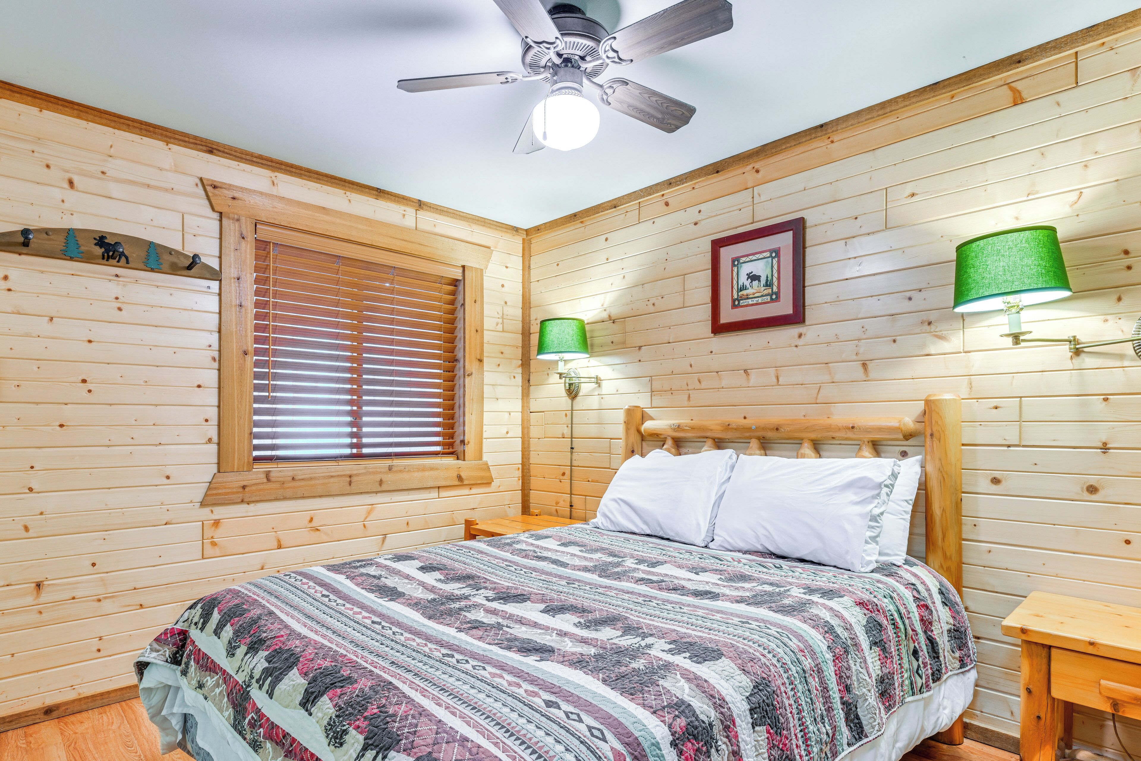 A wooden cabin-style bedroom with a bed, a ceiling fan, a window with blinds, and wall-mounted lamps.