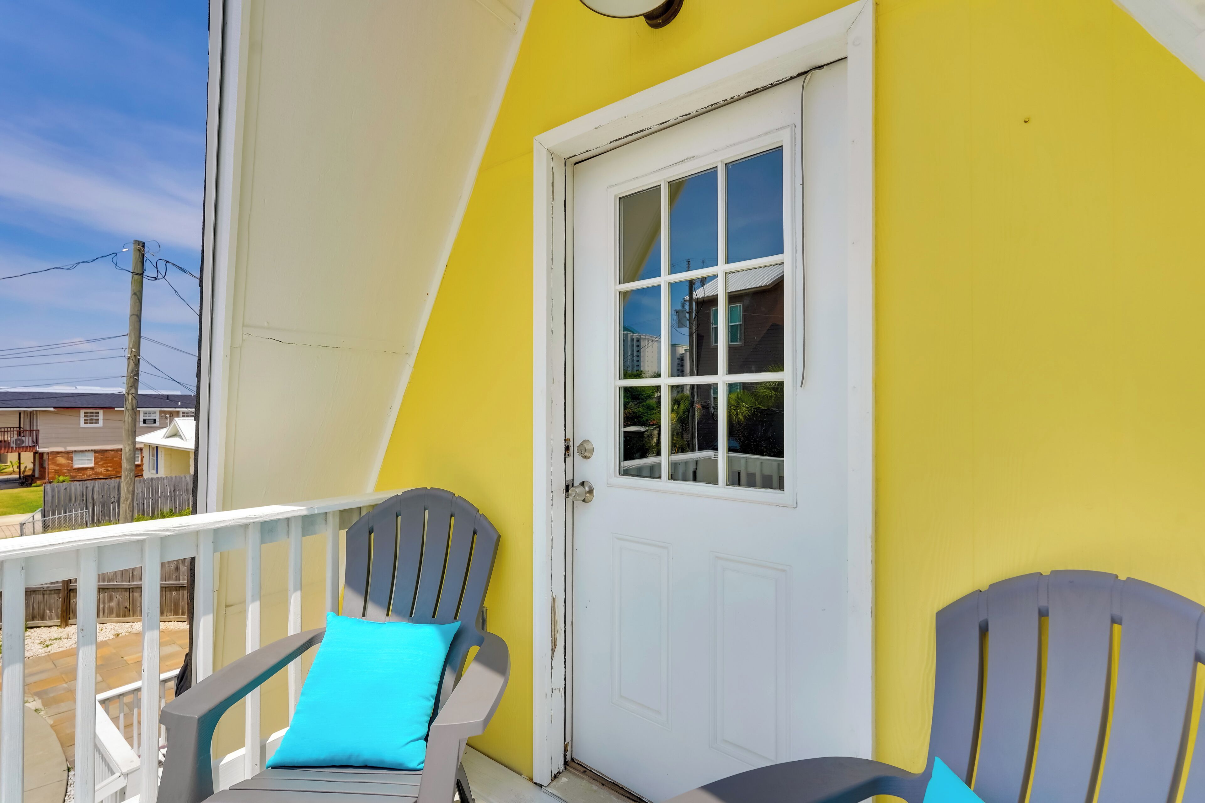 A yellow exterior wall with a white door and window, two gray Adirondack chairs with blue cushions, and a white railing on a balcony.