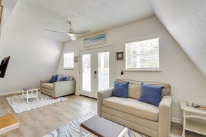 Ein geräumiges Wohnzimmer mit hoher Decke, einem Deckenventilator, einem beige farbenen Sofa mit blauen Kissen, einem weißen Couchtisch und einem weißen Beistelltisch.