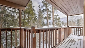 Un muelle de madera con barandilla, suelo cubierto de nieve y una vista de árboles y montañas al fondo.