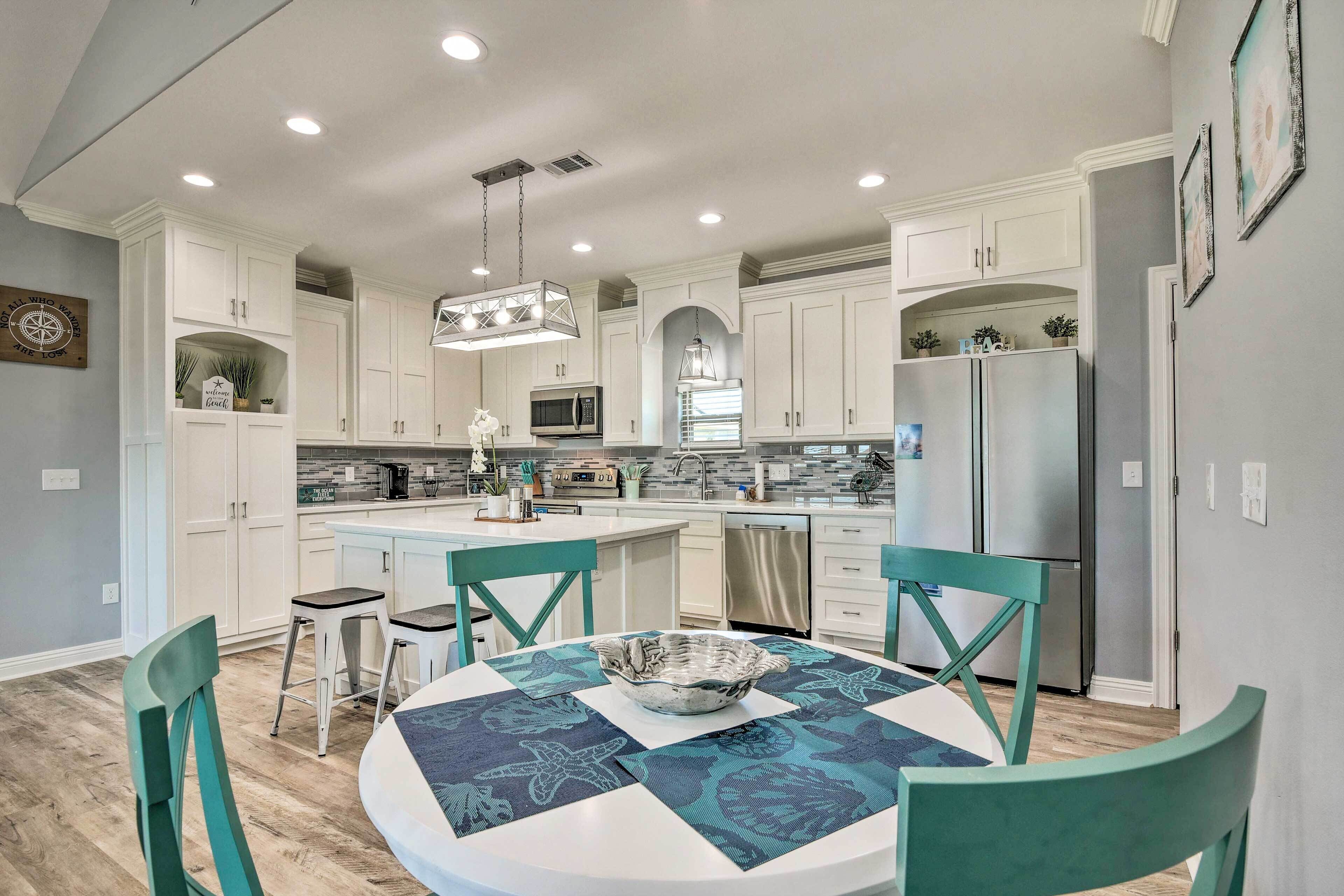 Une cuisine moderne avec des meubles blancs, un îlot central et des chaises turquoise disposées autour d’une table ronde sur laquelle se trouve un vase décoratif.
