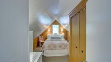 Un dormitorio con techo inclinado, una cama con ropa de cama estampada, una silla de madera y una ventana con luz natural.