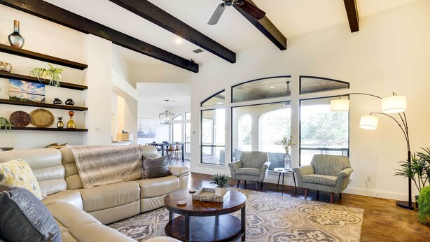 A spacious living room with a large sofa, a coffee table, and a patterned rug. There are shelves with decorative items and a large window providing natural light.