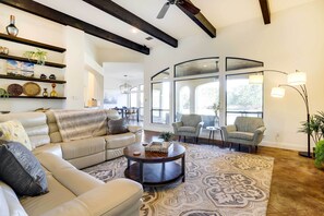 Un salon spacieux avec un grand canapé, une table basse et un tapis à motifs. On y trouve des étagères ornées d’objets décoratifs et une grande fenêtre qui laisse entrer la lumière naturelle.
