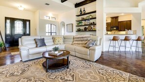 A spacious living room with a large sectional sofa, a coffee table, and patterned rugs. There is a kitchen area with bar stools and a countertop visible in the background.