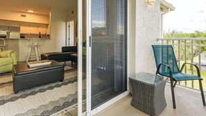 A living room with a sofa, a chair, and a small table. There is a sliding glass door leading to a balcony with a chair and a wicker table.