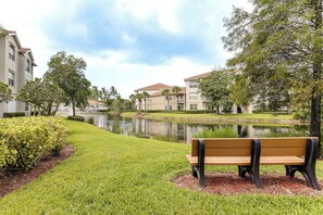 A bench in a park with a pond and buildings in the background.