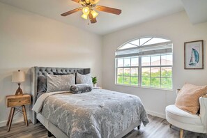 Une chambre à coucher avec une tête de lit capitonnée, une grande fenêtre offrant une belle vue, un ventilateur de plafond et une table de chevet avec une lampe.