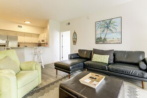 A living room with a sectional sofa, a green armchair, a coffee table, and a painting of a beach scene. - Work, Swim + Play in Ft Myers: Resort Condo Stay! (Fort Myers)
