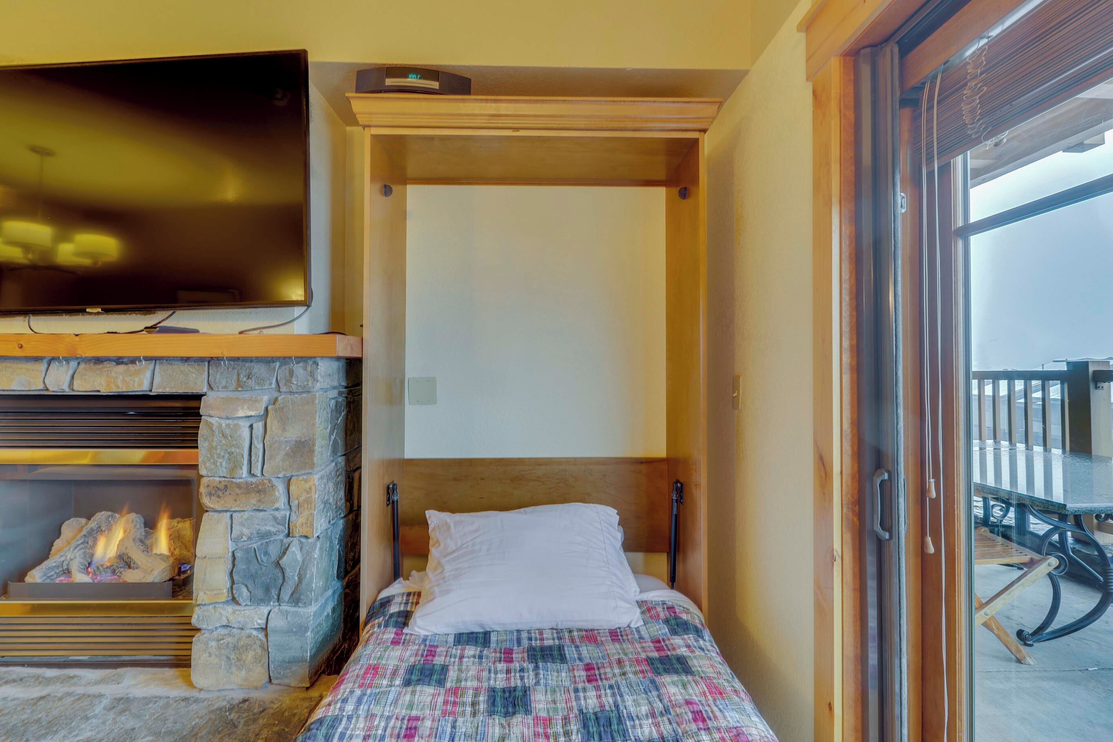 Un dormitorio con chimenea de piedra, una cama con colcha escocesa, un televisor sobre la chimenea y un ventanal corredero que da a un espacio exterior.