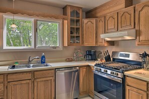 Een keuken met houten kasten, een roestvrijstalen vaatwasser, een fornuis en een raam met uitzicht op bomen.