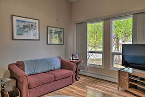 A living room with a red sofa, a television on a wooden stand, and large windows with a view of trees. - 8 Mi to Dtwn Achorage! Home w/ Pet-friendly Yard (Anchorage)
