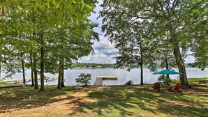 A lakeside view with a dock, picnic table, and chairs under a tree. - 'the B Haus' Lakefront Retreat w/ Boat Ramp! (Talladega)