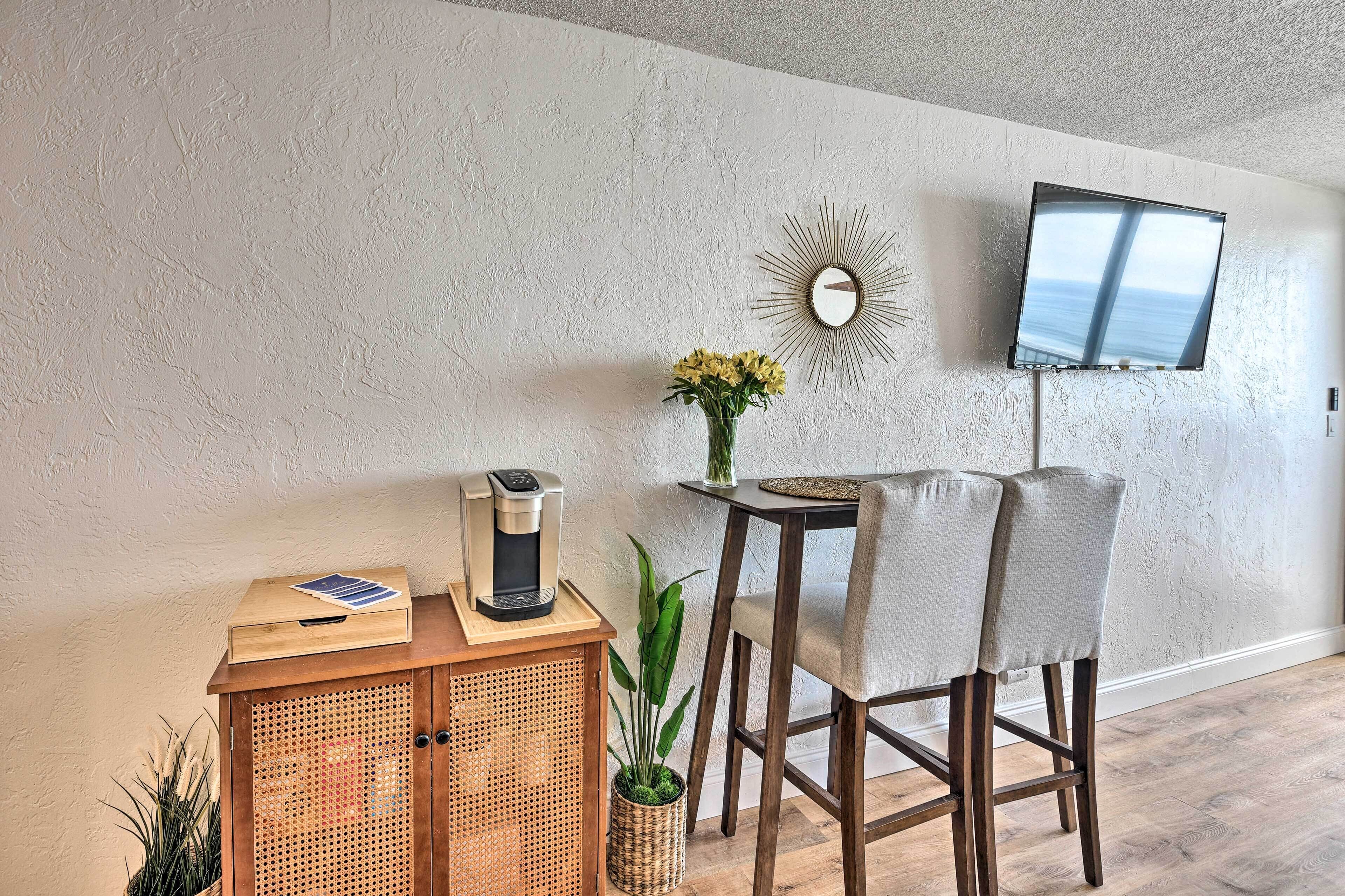 Ett modernt vardagsrum med en kaffebryggare på en träkonsol, ett litet bord med en vas med blommor och en tv som är monterad på väggen.
