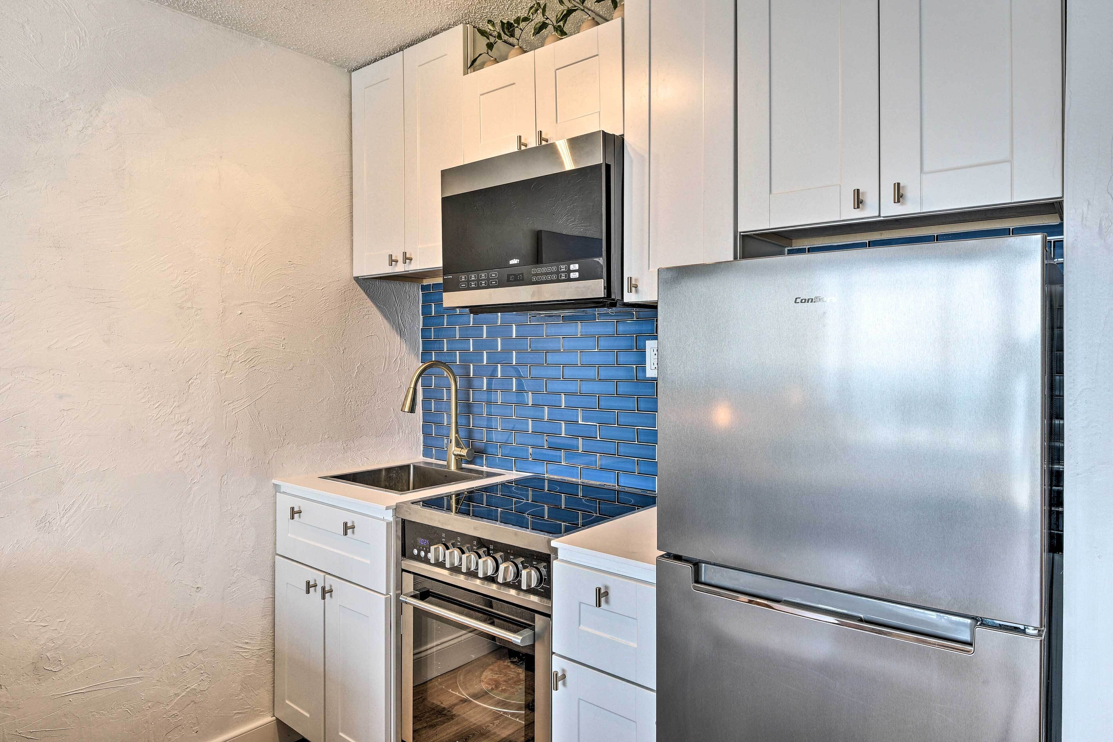 A compact kitchen with white cabinets, a stainless steel refrigerator, a built-in oven, and a blue-tiled backsplash.