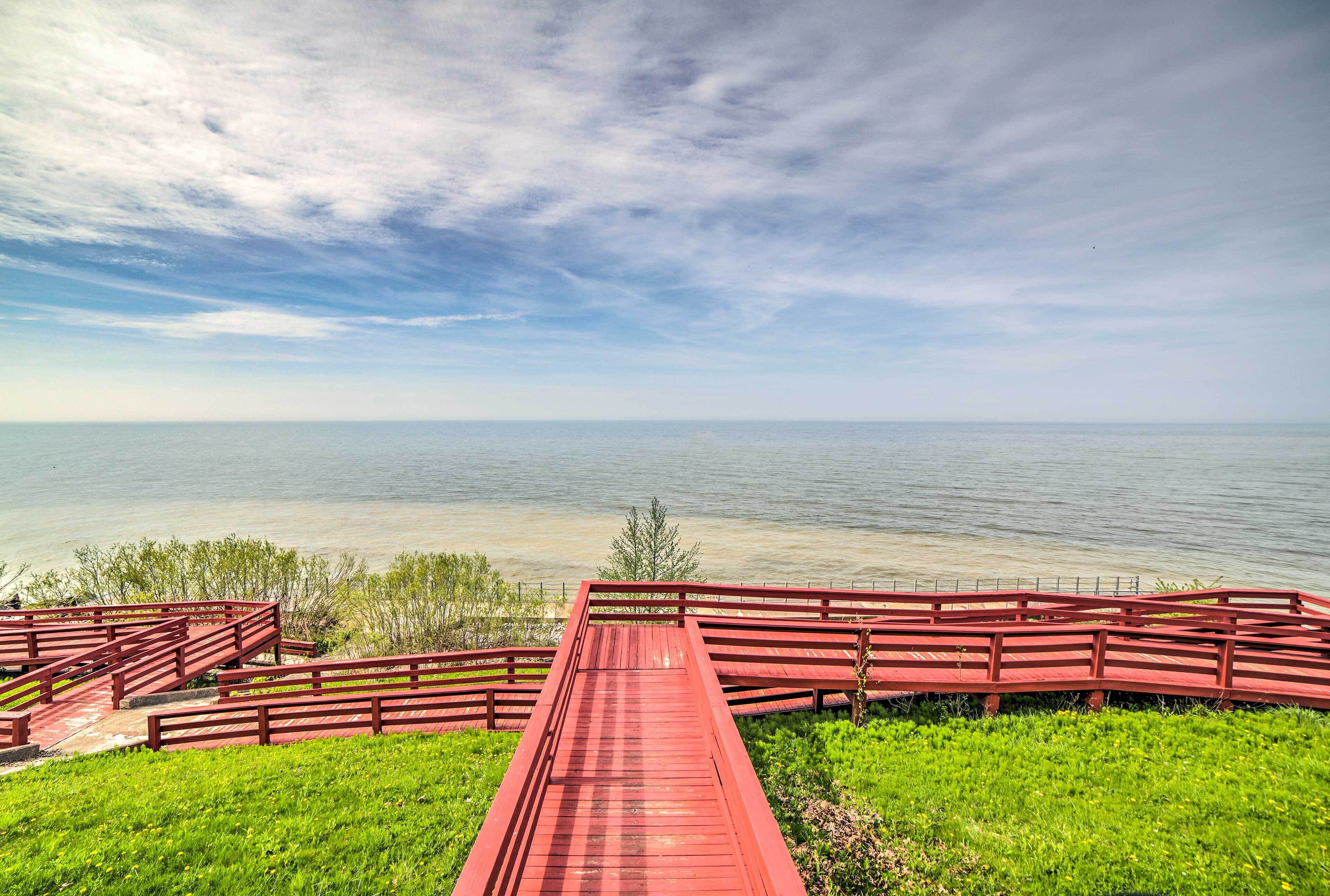 Eine rote Holzbrücke führt zu einer grünen Wiese mit Blick auf ruhiges Wasser unter einem bewölkten Himmel.