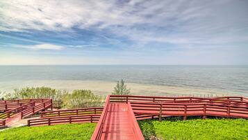 A red wooden bridge leading to a grassy area with a view of a calm body of water under a cloudy sky.