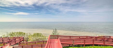 A red wooden bridge leading to a grassy area with a view of a calm body of water under a cloudy sky.