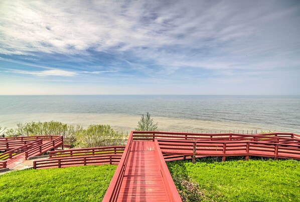 A red wooden bridge leading to a grassy area with a view of a calm body of water under a cloudy sky.