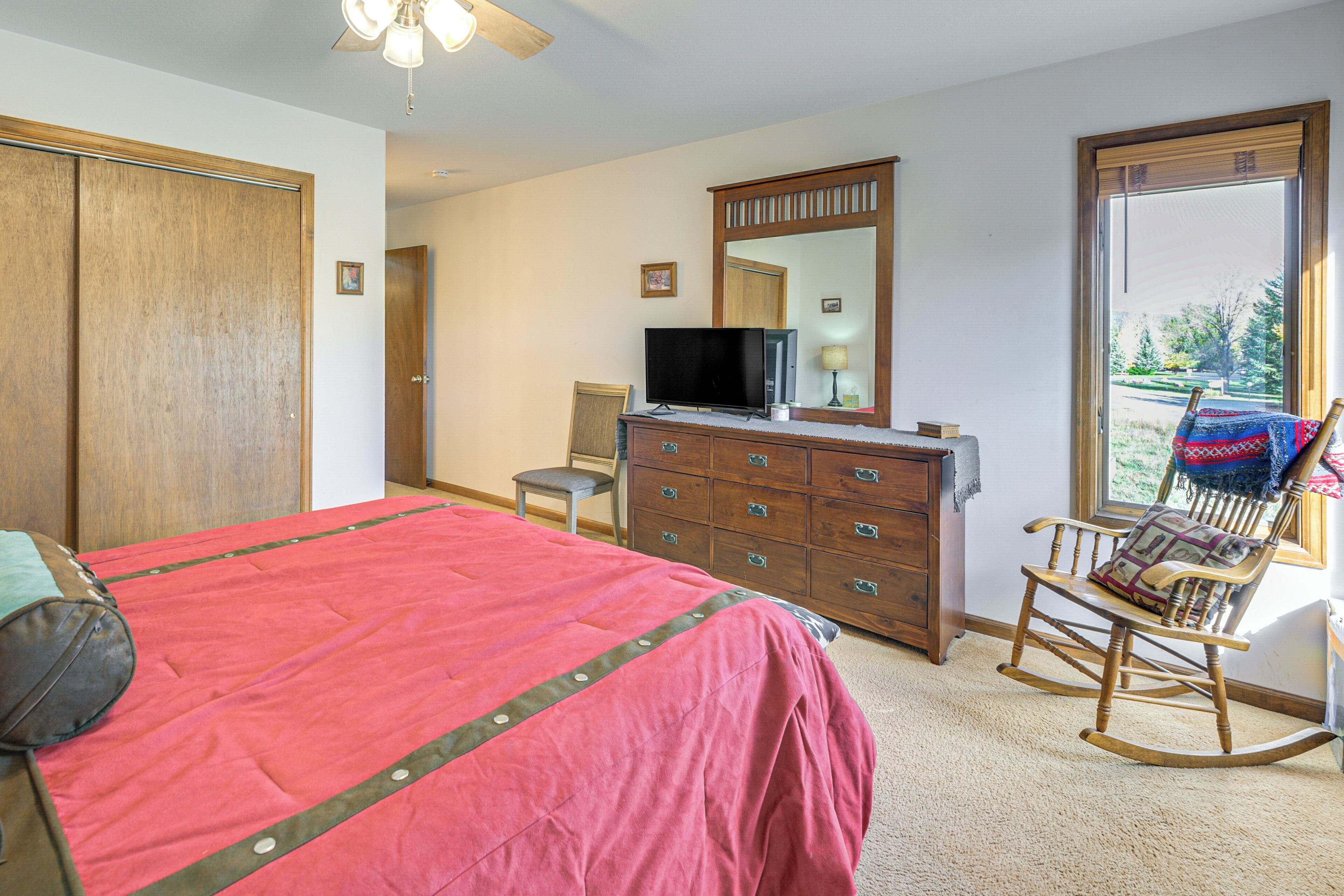 A bedroom with a bed, dresser, television, rocking chair, and a large window.