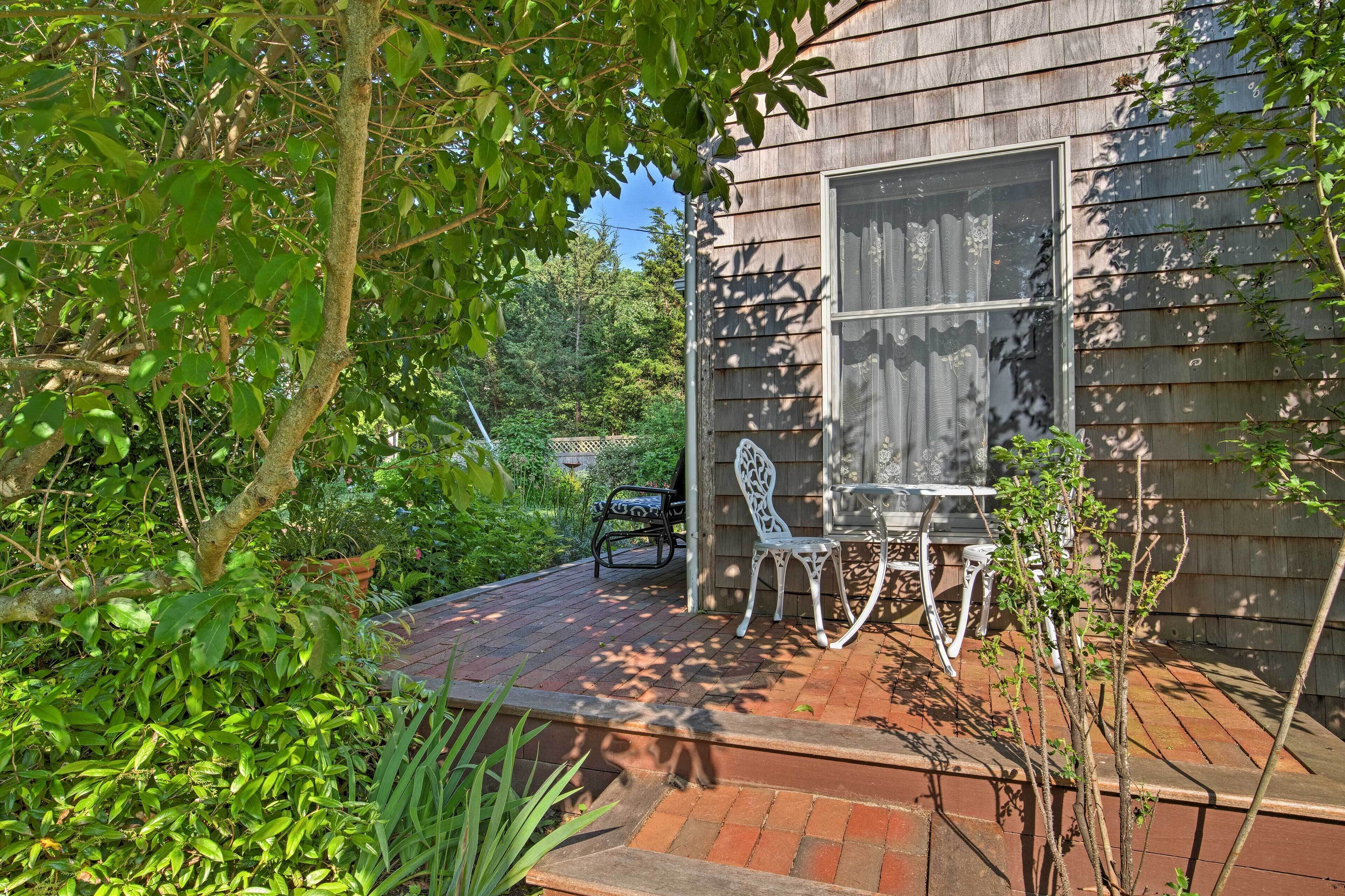 A wooden house with a brick patio, white outdoor furniture, and a window with curtains.