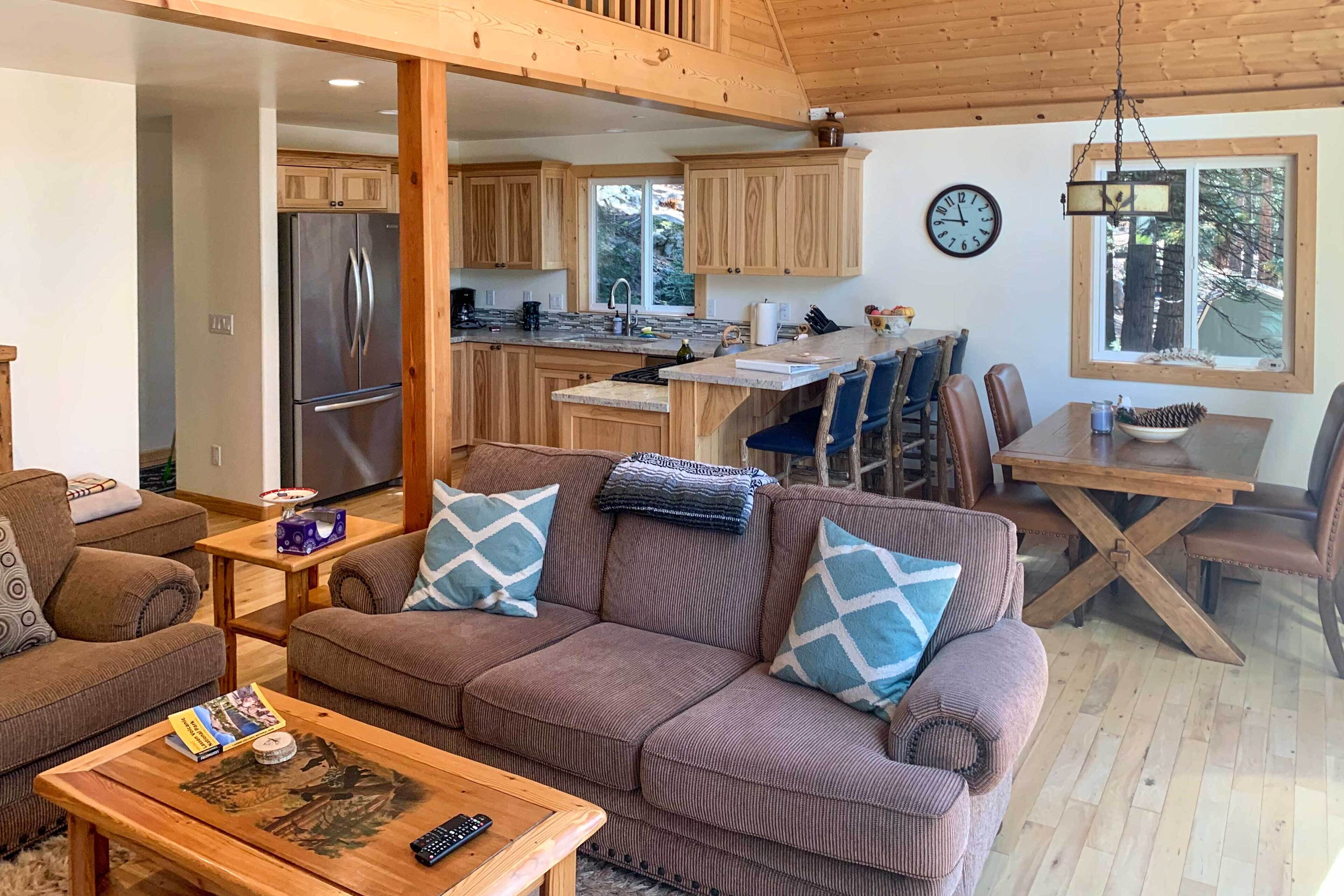 Ein gemütliches Wohnzimmer mit einer braunen Couch, einem Holztisch und einem Essbereich mit Tisch und Stühlen. Im Hintergrund ist eine Küche mit Holzschränken und einem Kühlschrank erkennbar.
