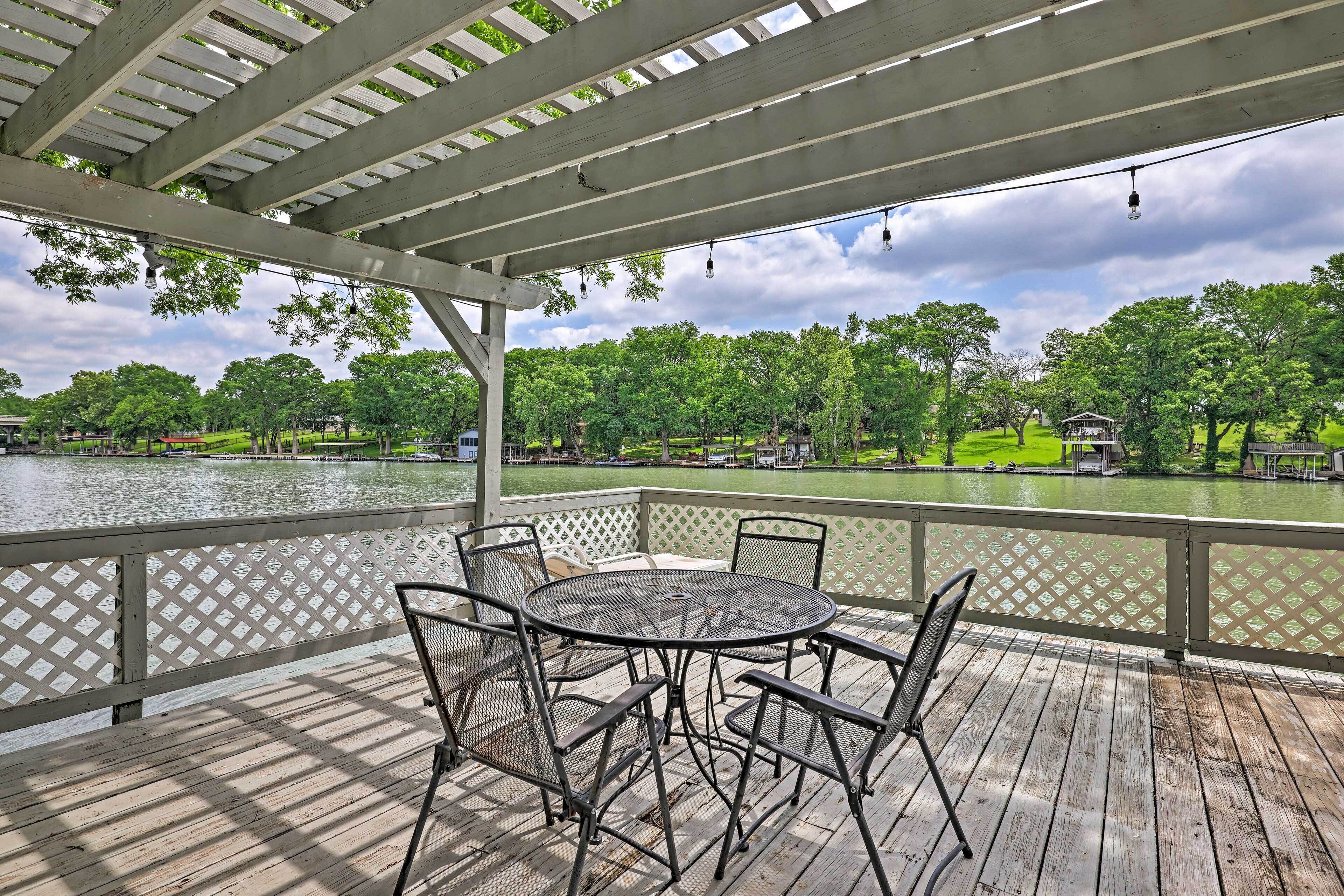 A covered outdoor area with a table and chairs, overlooking a lake with trees and a house in the background.