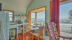 A cozy kitchen with wooden flooring, a dining table with chairs, a refrigerator, and a large window offering a view of the ocean.