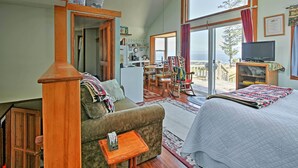 A cozy living room with a sofa, a small table, and a bed. There is a kitchen area with a refrigerator and a dining table. The room has large windows with curtains and a view of the outdoors.