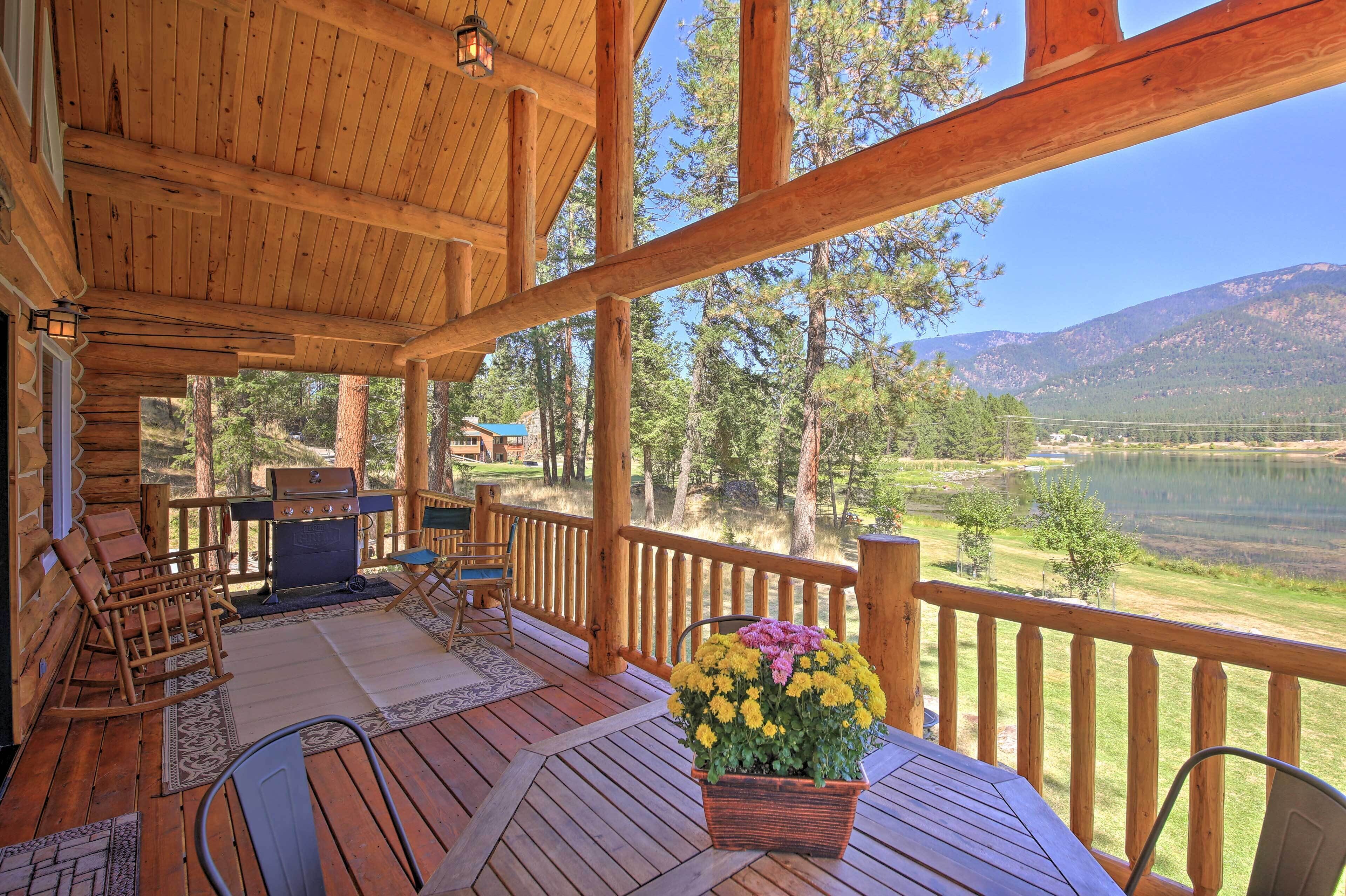 Una terraza de madera con mobiliario exterior, una parrilla de barbacoa y vistas a un lago y montañas.